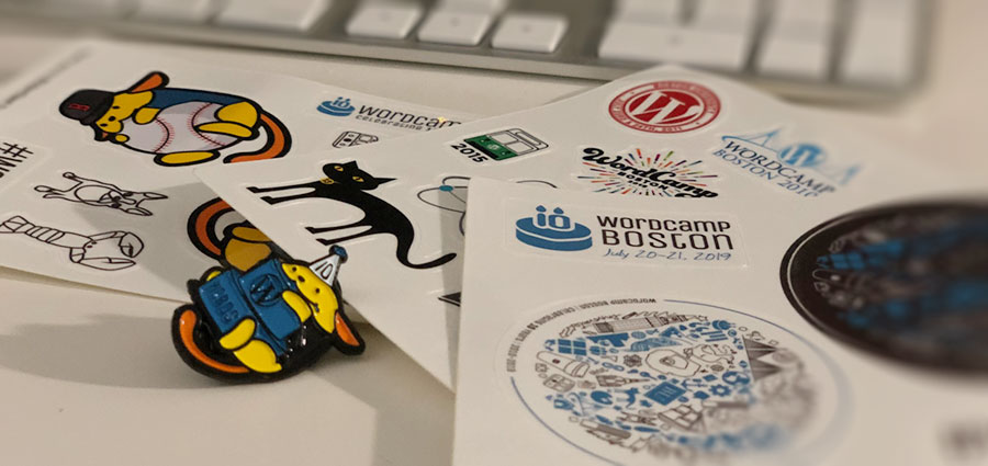 Sticker sheets from WordCamp Boston 2019 showing current and past logos, arranged on desk with keyboard and Wapuu pins
